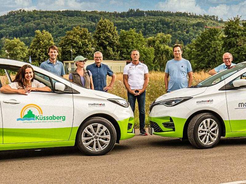 Eine Gruppe von Menschen steht lächelnd neben zwei mit Werbeaufschriften versehenen Elektroautos im Freien.