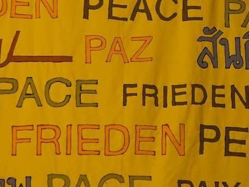 Das Bild zeigt ein gelbes Banner mit dem Wort "Frieden" in verschiedenen Sprachen und Schriftarten darauf geschrieben.