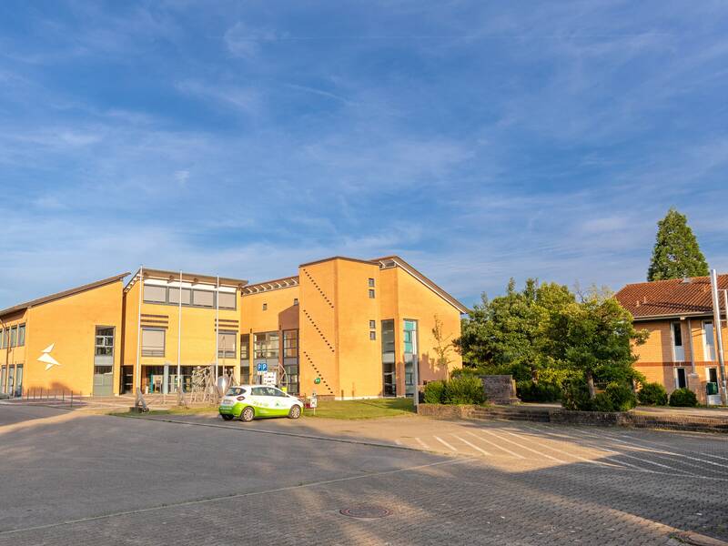 Eine breite Straße führt zu einem modernen Gebäudekomplex unter einem blauen Himmel, begleitet von einem grün-weißen Auto und umgeben von kleinen Bäumen.