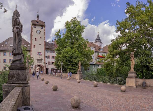 eine Brücke mit Steinstatuen auf dem Geländer und große runde Steinkugeln als abgrenzung zwischen Fußgängern und Autos, am Ende der Brücke steht ein Stadttor mit Uhr und angrenzenden Häusern