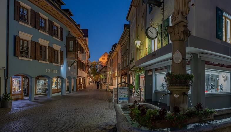 eine gepflasterte Fußgängerzone in der Abenddämmerung rechts vorne ein Stadtbrunnen, links und recht ist die Zone von geschlossenen Geschäften gezäumt