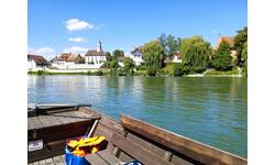 Blick von einer Holzfähre auf dem Rhein mit Blick über das Wasser in Richtung rheinabwärts auf das deutsche Rheinufer des Ortsteils Kadelburg mit Blick auf die Kirche und Wohngebäude bei blauem Himmel