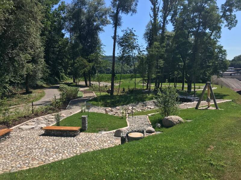 ein gepflasterter Weg und unterschiedliche Kies- und Stein- und Holzwege schlängeln sich durch eine grüne Parkanlage mit Sitzbänken und einer Bepflanzung, im Hintergrund blauer Himmel und Bäume.