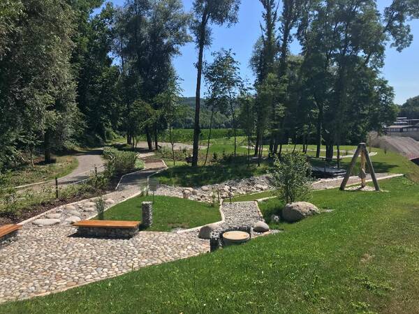 ein gepflasterter Weg und unterschiedliche Kies- und Stein- und Holzwege schlängeln sich durch eine grüne Parkanlage mit Sitzbänken und einer Bepflanzung, im Hintergrund blauer Himmel und Bäume.