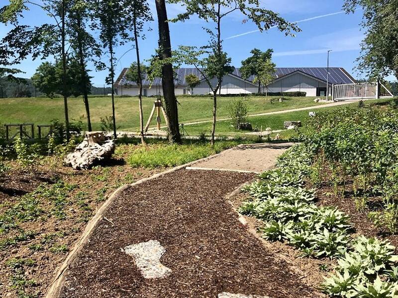 ein Weg aus Rindenmulch mit zwei großen geformten Fußabdrücken als Steinbett eingelassen in den Weg, welcher an beiden Seiten grün bepflanzt ist.  In der Fortführung des Weges folgt ein mit Sand gefüllter Wegabschnitt. Blauer Himmel und Bäume im Hintergrund des Bildes. 