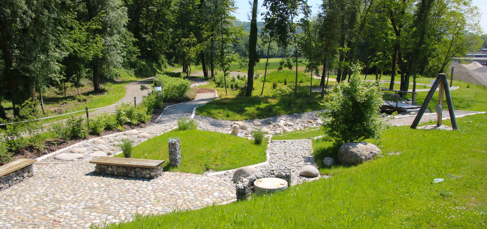 ein gepflasterter Weg und unterschiedliche Kies- und Stein- und Holzwege schlängeln sich durch eine grüne Parkanlage mit Sitzbänken und einer Bepflanzung, im Hintergrund führt ein Weg weiter entlang von Bäumen.