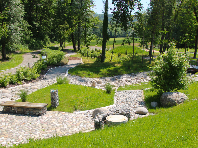 ein gepflasterter Weg und unterschiedliche Kies- und Stein- und Holzwege schlängeln sich durch eine grüne Parkanlage mit Sitzbänken und einer Bepflanzung, im Hintergrund führt ein Weg weiter entlang von Bäumen.