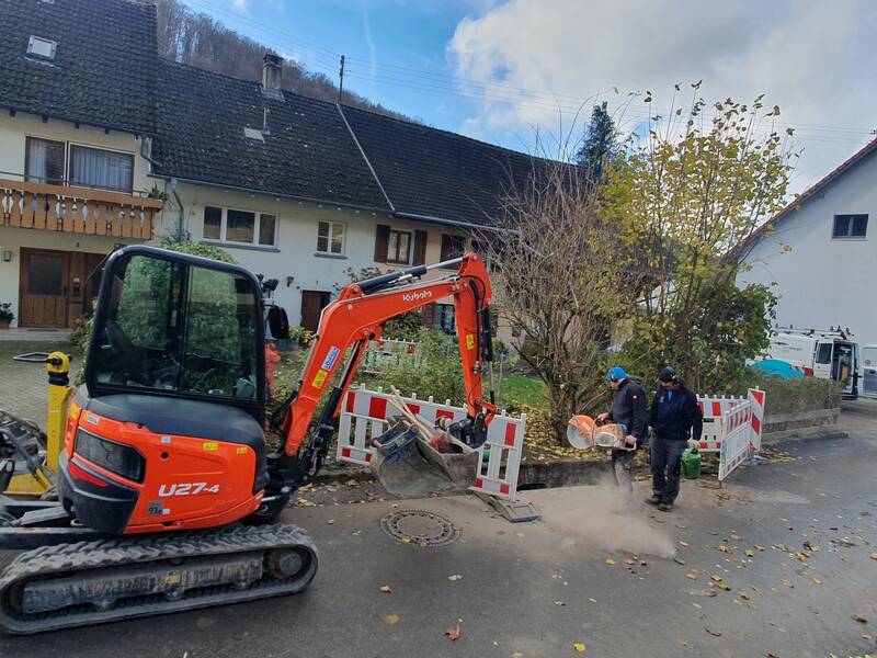 Zwei Arbeiter führen Bauarbeiten an einem Weg neben einem Wohnhaus durch, mit Absperrungen und einem roten Bagger links im Hintergrund