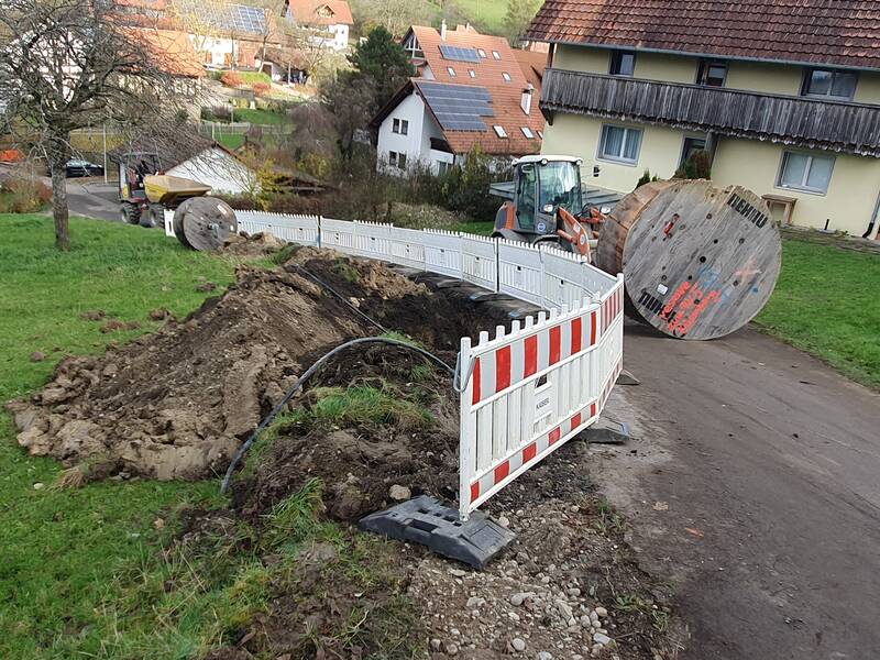 Auf einem Feldweg in einem Dorf wird ein Graben für Bauarbeiten ausgehoben, gesichert durch Absperrungen, während ein Bagger und große Kabeltrommeln sichtbar sind.