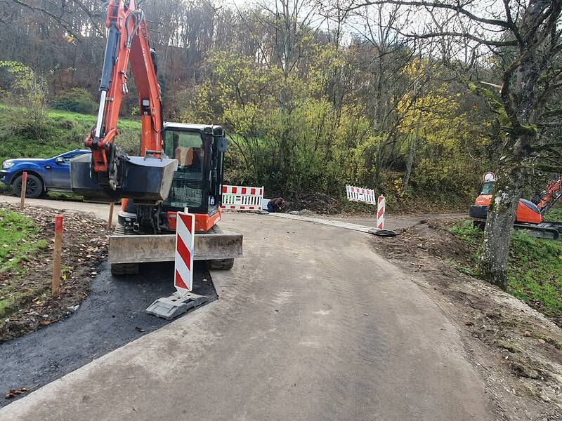 Eine Baustelle auf einem schmalen Weg mit zwei roten Baggern, Absperrungen und einem blauen Auto im Hintergrund, umgeben von Bäumen und Wiesen.