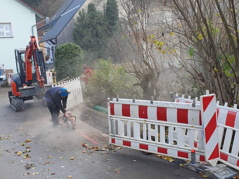 Ein Arbeiter verwendet ein Schneidwerkzeug auf einer Straße neben einer Baustelle mit Absperrzäunen und einem Bagger.