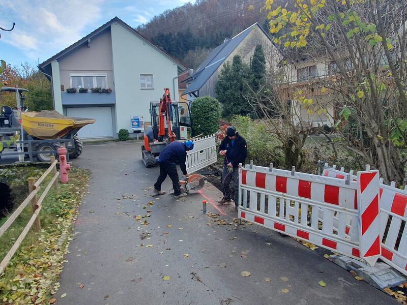 Zwei Arbeiter führen Bauarbeiten an einem Weg neben einem Wohnhaus durch, mit Absperrungen und einem Bagger im Hintergrund.