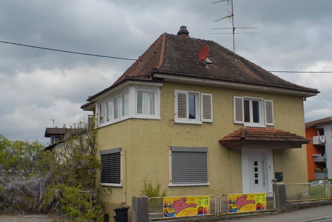 Ein gelbes Einfamilienhaus mit geschlossenen Fensterläden und einer roten Satellitenschüssel auf dem Dach, vor einem bewölkten Himmel.