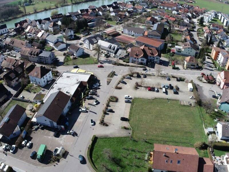 Luftbild des Ortskerns Kadelburg, Kreuzungsbereich, Straßenverlauf der Ortsdurchfahrtsstraße Hauptstraße mit Kreuzung in die Kirchstraße und Deckwiesenstraße. Ansicht von oben auf umliegende Wohnhäuser, das Apotheken- und Sparkassengebäude, zentral mittig das große EDEKA Einkaufsgebäude und Park- und Grünflächen sowie links im Bild ein Arztpraxisgebäude. Im Hintergrund sieht man den Rheinverlauf.