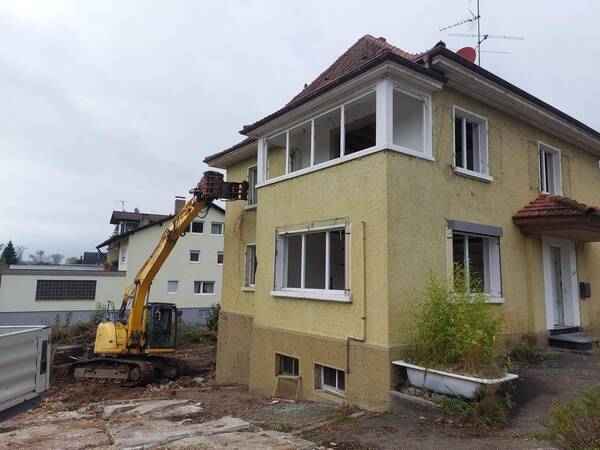 Ein gelbes altes zweistöckiges Gebäude, die Glasscheiben fehlen, ein Bagger beginnt mit Abrissarbeiten und hat die Schaufel oben links an der Hauswand angesetzt, vor einem bewölkten Himmel.