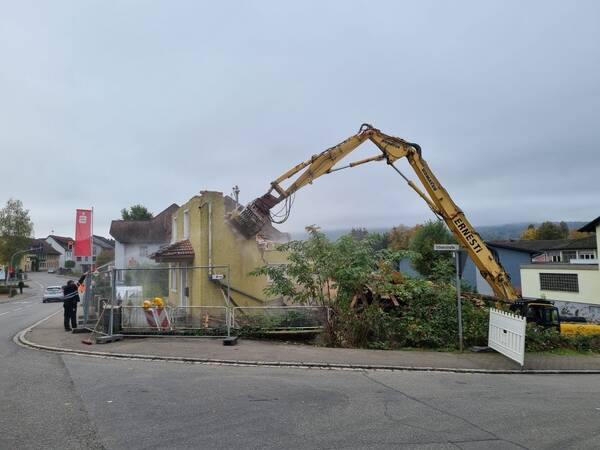 Ein altes zur Hälfte abgerissenes Gebäude, viele große Mauertrümmer, ein Bagger bei den Abrissarbeiten, ein Bauarbeiter hinter einem Bauzaun stehend, bei bewölktem Himmel.