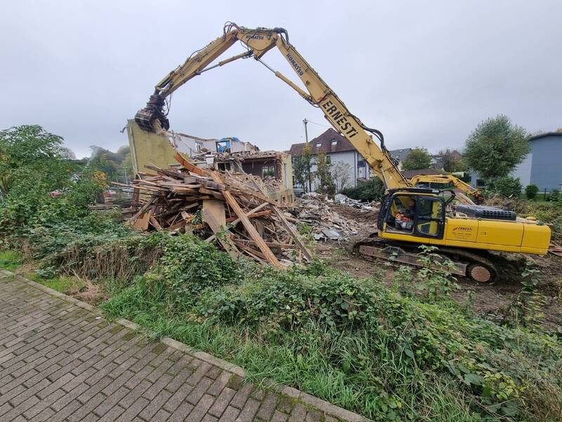 ein Bagger bei den Abrissarbeiten, im Vordergrund ein großer Haufen Abrißholz, im Hintergrund Mauerrest eines Gebäude, viele große Mauertrümmer, bei bewölktem Himmel.