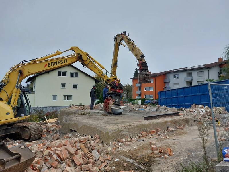 Auf einer Baustelle hebt ein gelber Bagger Ziegelsteine, während im Hintergrund Wohngebäude sichtbar sind.