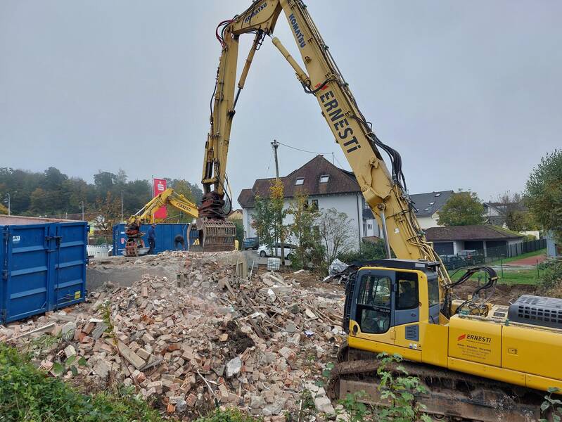 Ein gelber Bagger räumt Schutt auf einer Baustelle ab, während ein weiterer Bagger im Hintergrund sichtbar ist.