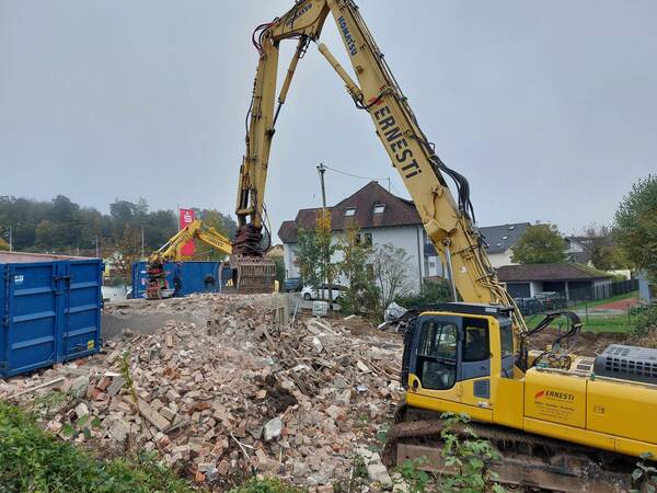 Eine Baustelle mit einem großen gelben Bagger, der auf einem Haufen Ziegeltrümmer arbeitet, während blaue Container im Hintergrund zu sehen sind.