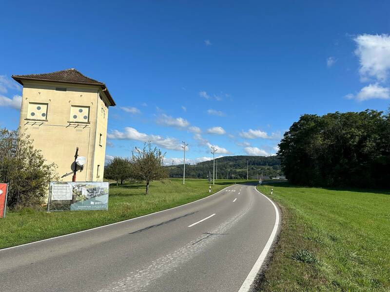 Blick auf den Straßenverlauf Ortsausgang Rheinheim in Richtung Dangstetten mit Blick auf die defekte Asphaltdecke, links eine Travostation, davor am Wegesrand ein Werbebanner, an beiden Seiten Grünlandschaft, blauer Himmel.