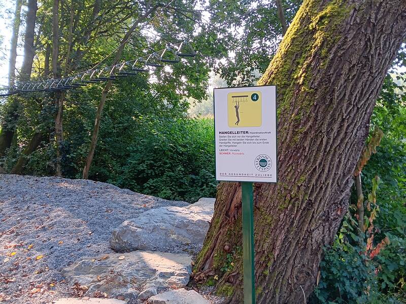 Das Bild zeigt einen Kletterparcours im Wald mit einer Hangeleiter und einem erklärenden Schild davor.