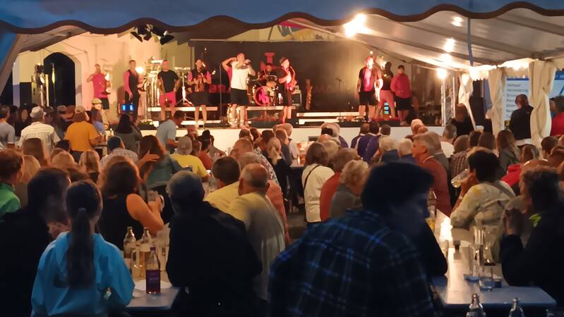 Im Festzelt, im Vordergrund sieht man viele Besucher an Biergarnituren sitzen auf der Bühne sielt eine Band mit 11 Musikern