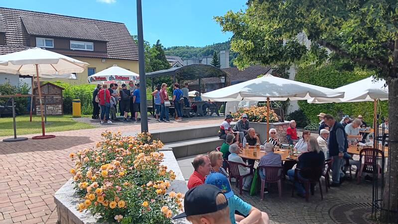 ein Dorfplatz mit zwei Ebenen, eine Gruppe von Menschen sitzt unter Sonnenschirmen an Tischen auf der niedrigeren Ebene, zwei hohe Stufen, auch als Sitzplatz geeignet führen zur oberen Ebene, hier steht eine weitere Gruppe.