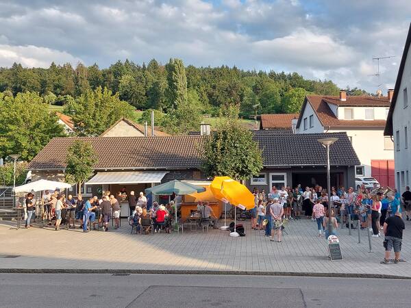 Blick von der anderen Straßenseite auf eine Dorfplatz mit vielen Leuten, Stehtischen und Sitzgelegenheiten