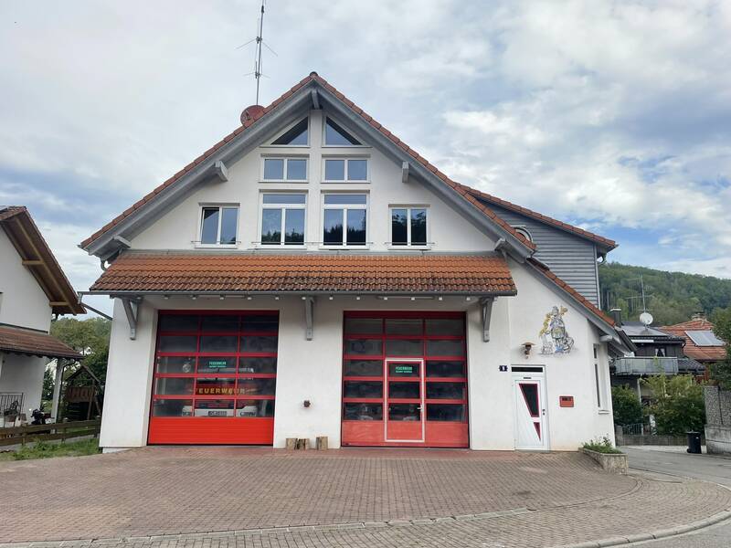 Das Bild zeigt ein zweistöckiges Feuerwehrhaus mit weißen Wänden, einem roten Dach und zwei großen roten Garagentoren, vor einem bewölkten Himmel.