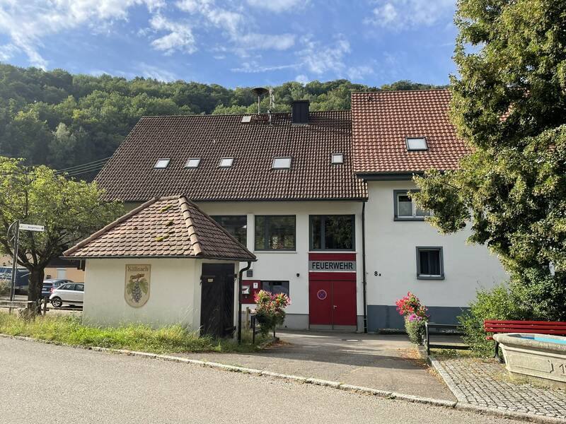 Feuerwehrhaus mit roten Tor und einem kleinen Häuschen davor mit dem Küßnacher Wappen. Im Hinter ist der Wald und blauer Himmel zu sehen 