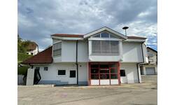 Das Bild zeigt ein modernes, zweigeschossiges Feuerwehrhaus mit einem roten Garagentor und einem sichtbaren Feuerwehrschild, umgeben von einem Parkplatz und anderen Gebäuden unter einem bewölkten Himmel.