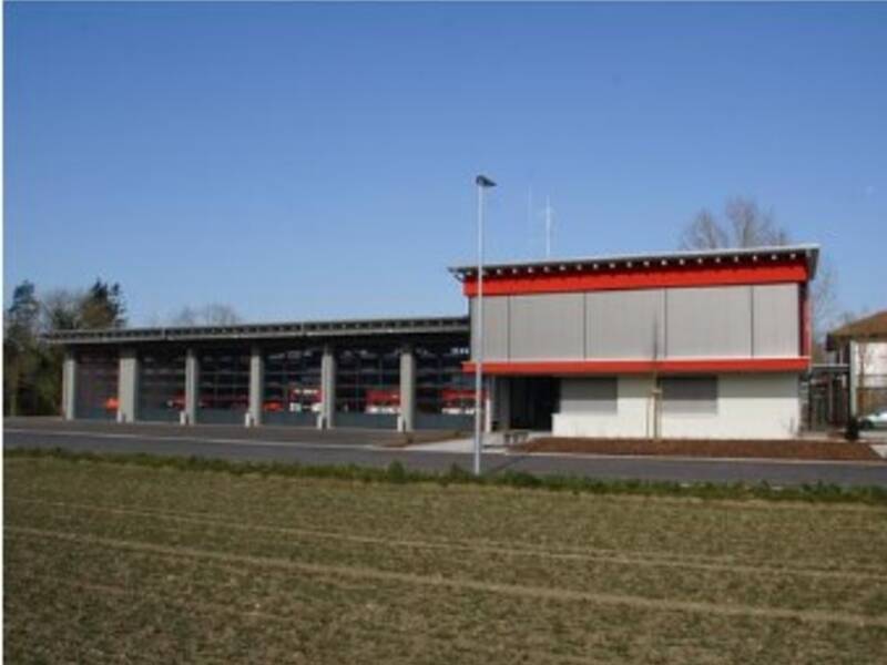 Ein modernes, eingeschossiges Feuerwehrhaus mit rotem Dachrand und mehreren Garagentoren unter einem blauen Himmel.