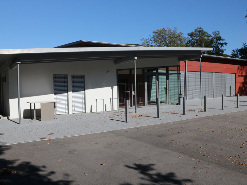 Ein modernes Gebäude mit flachem Dach, großen Fenstern und einer betonierten Vorplatz ist bei sonnigem Wetter zu sehen.