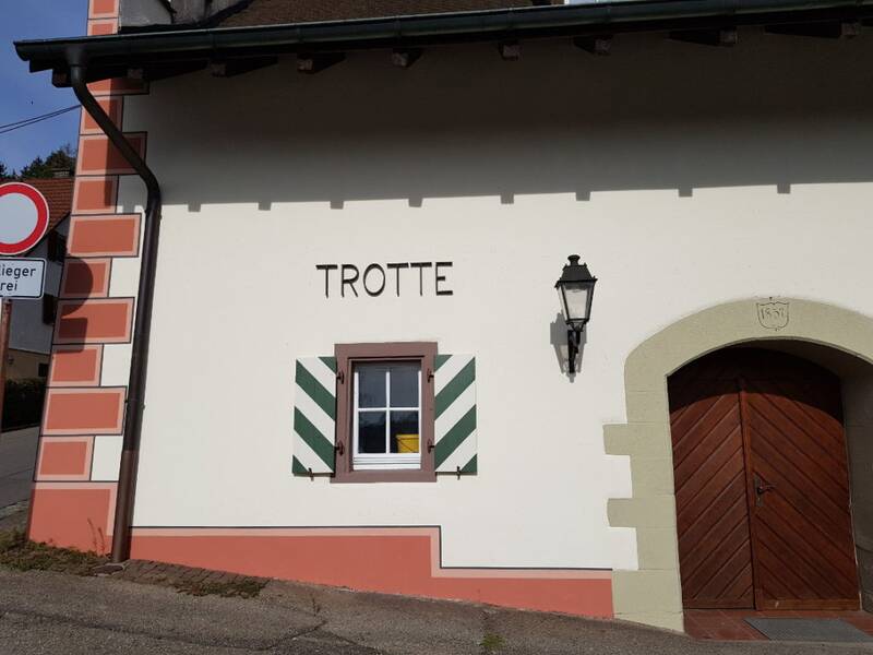 Auf dem Bild ist die Fassade eines traditionellen Gebäudes mit dem Schriftzug "TROTTE" und der Jahreszahl "1857", einer Holztür und einem Fenster mit grünen Fensterläden zu sehen.