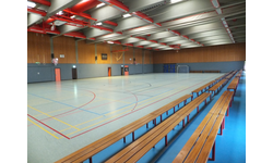 Große Sporthalle mit Basketballkörben, farbigen Spielfeldmarkierungen auf dem Boden und einer Sitztribüne auf einer Seite.