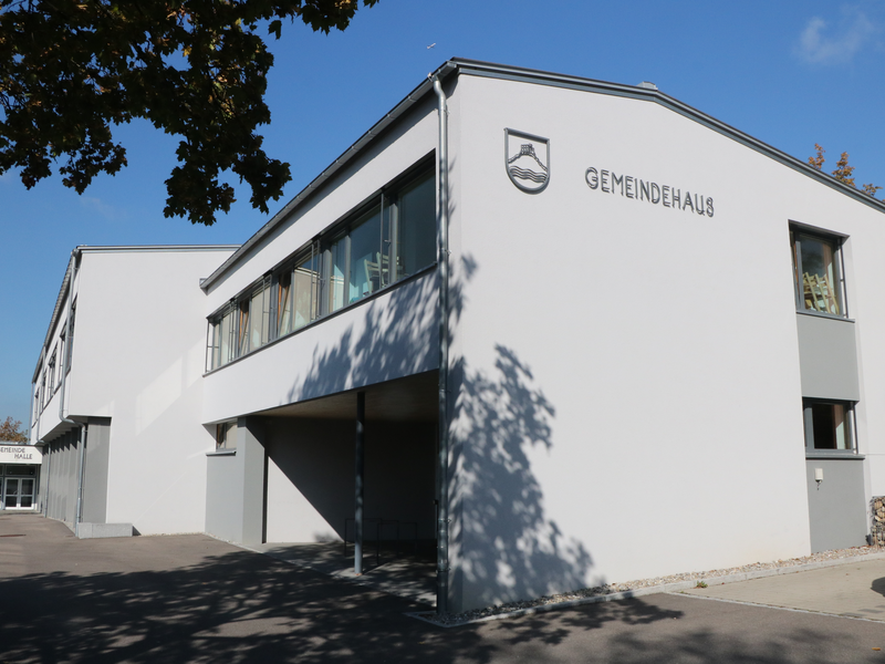 Modernes weißes Gemeindehaus mit großen Fenstern unter einem blauen Himmel, umgeben von Bäumen und einem gepflasterten Vorplatz. Auf der Fassade sind das Gemeindewappen und ein Schriftzug Gemeindehaus angebracht. 