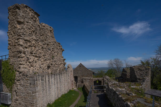 Eine alte, teils überwucherte Burgruine unter klarem, blauem Himmel mit Blick auf eine weite Landschaft im Hintergrund.