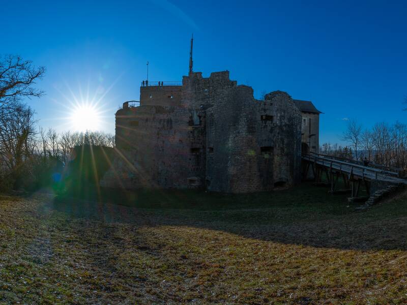 Eine alte Burgruine steht in einer bewaldeten Landschaft unter einem strahlend blauen Himmel mit der Sonne im Hintergrund.