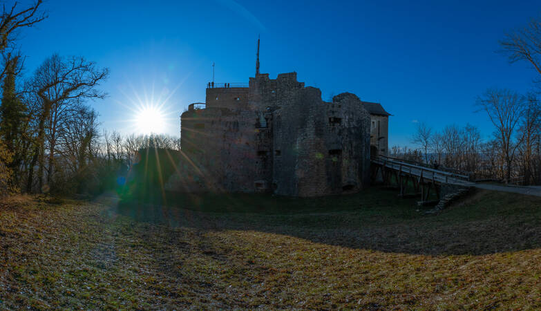 Eine alte Burgruine steht in einer bewaldeten Landschaft unter einem strahlend blauen Himmel mit der Sonne im Hintergrund.