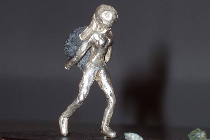 eine kleine Skulptur einer Frau aus einem metallischen Material, die auf dem Rücken im Verhältnis einen sehr großen Stein trägt