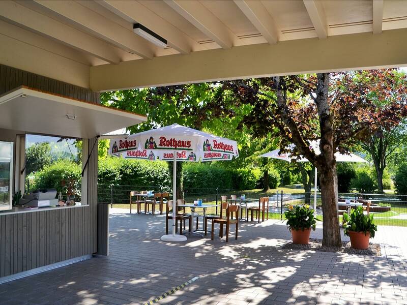 Ein Kiosk mit überdachten Sitzmöglichkeiten, Sonnenschirmen und einem Baum inmitten einer grünen Umgebung.