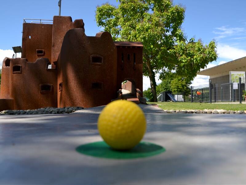 Eine gelbe Minigolfball-Kugel liegt auf einem Golfrasen vor einem rotbraunen Gebäude mit Bäumen im Hintergrund.