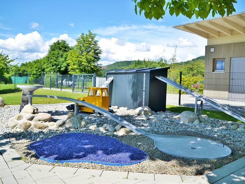 Eine moderne Minigolfstation aus Metall, umgeben von Kieselsteinen und einem Haus im Hintergrund.