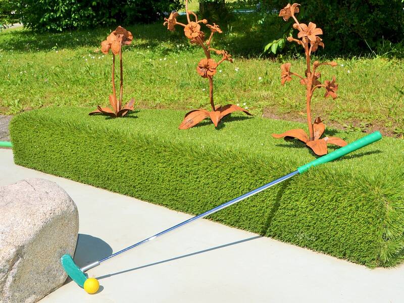 Ein Mini-Golf-Schläger mit grünem Griff und gelbem Ball liegt auf einer Betonfläche vor einem künstlichen Rasen, der von metallischen Blumen geschmückt ist.