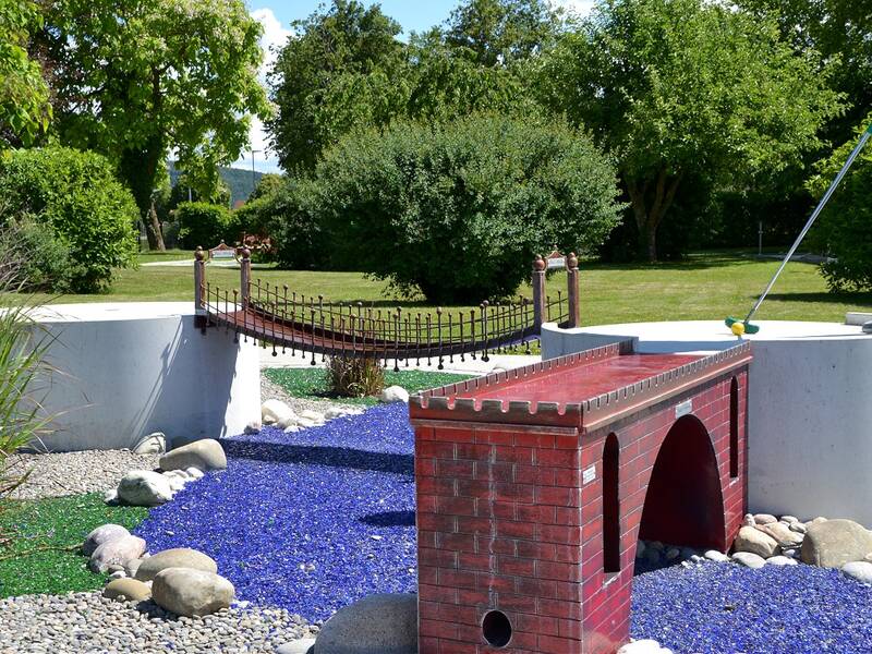 Ein Minigolfplatz mit einer roten Ziegelbrücke und blauem Kies, auf dem eine Person mit einem Golfschläger steht, umgeben von grünen Büschen und Bäumen.