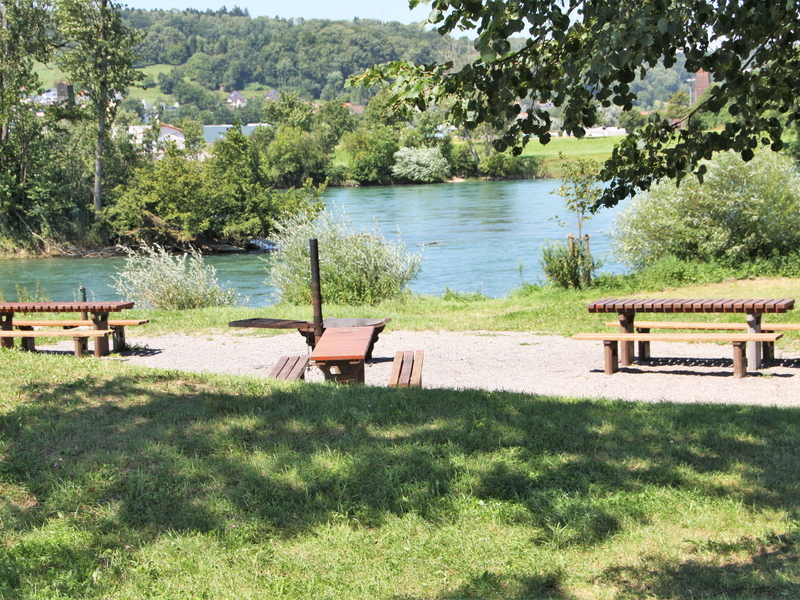 Ein idyllischer Rast- und Grillplatz mit mehreren Holzbänken und einem Tisch am Ufer eines Flusses umgeben von Grünflächen und Bäumen an einem sonnigen Tag.