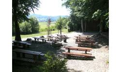 Ein idyllischer Rast- und Grillplatz am Waldrand auf einer Waldlichtung mit mehreren Holzbänken und Tischen im Schatten großer Bäume mit Blick auf eine grüne Landschaft.