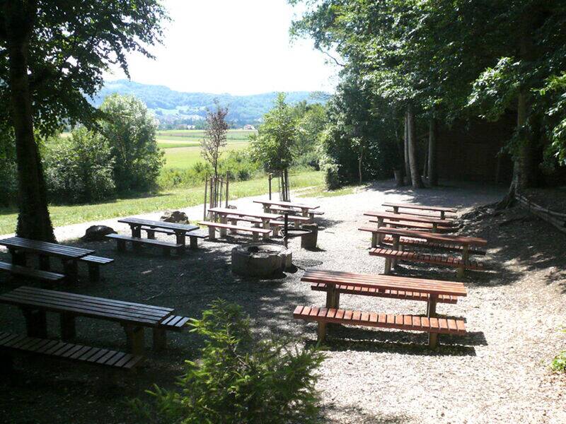 Ein idyllischer Rast- und Grillplatz am Waldrand auf einer Waldlichtung mit mehreren Holzbänken und Tischen im Schatten großer Bäume mit Blick auf eine grüne Landschaft.