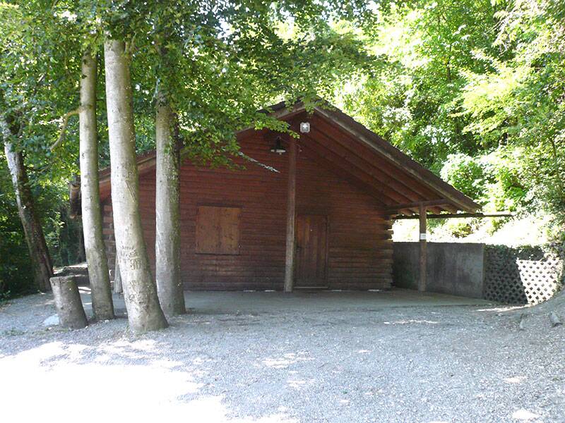 eine große Holzhütte umgeben von lichtdurchfluteten Bäumen, am Waldrand gelegen, im Vordergrund ein großer Rastplatz angedeutet. 